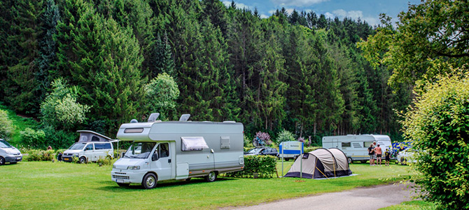 Mehrere Wohnmobile und Zelte sind auf einer Wiese in der Nähe eines Waldes aufgestellt, in der Nähe stehen Menschen unter einem teilweise bewölkten Himmel.