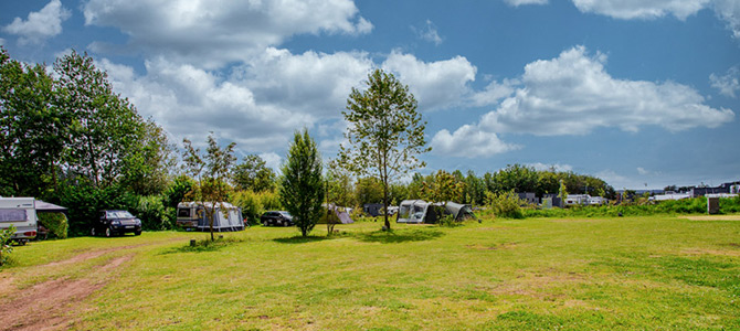 Offener, grasbewachsener Campingplatz mit mehreren Zelten, Wohnwagen und geparkten Autos, umgeben von Bäumen unter einem teilweise bewölkten Himmel.
