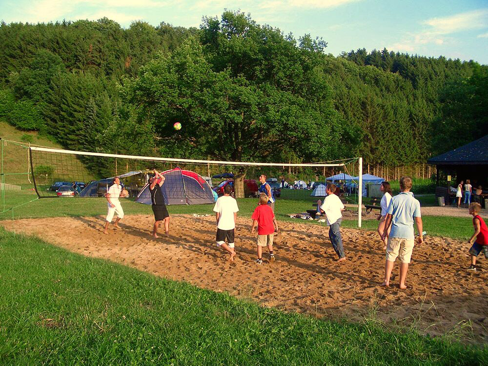 Eine Gruppe von Menschen spielt Volleyball auf einem Sandplatz im Freien, umgeben von Zelten, Gras und Bäumen.