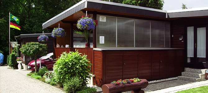 Eine moderne, braune Holzhütte mit hängenden Blumenkörben, Topfpflanzen und einem Fahnenmast mit zwei Flaggen in einem begrünten Hof.