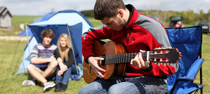 Een jonge man zit op een blauw campingstoeltje en speelt op een akoestische gitaar, met twee mensen die op het gras zitten en een tent op de achtergrond.