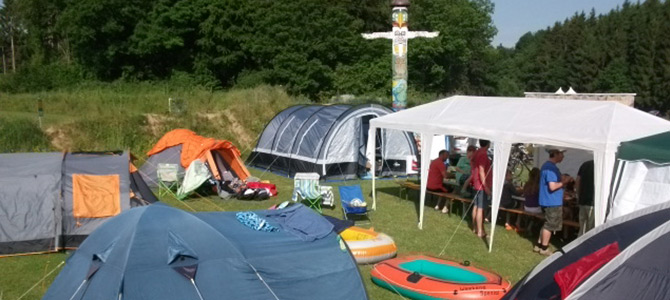 Verschillende tenten en een opblaasboot staan opgesteld op gras naast bomen, met mensen die onder een witte luifel zitten en een beschilderde totempaal zichtbaar op de achtergrond.