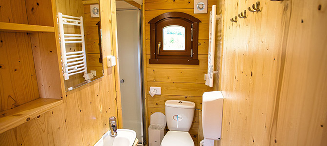 Kleines Holzbadezimmer mit Toilette, Waschbecken, Spiegel, Duschkabine, Handtuchhalter, Haken und einem kleinen Fenster, das Tageslicht hereinlässt.