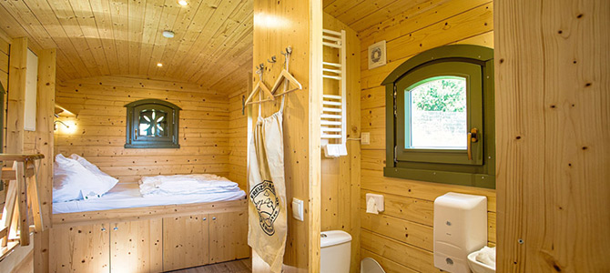 Kompakte Holzkabine mit einem sauber gemachten Bett, kleinen Fenstern, Kleiderhaken und einem angrenzenden Badezimmer mit Toilette und Handtuchhalter.