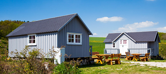 Zwei kleine graue Holzhütten mit weißer Verkleidung stehen auf einer Wiese, in der Nähe stehen Holztische und -stühle unter einem klaren blauen Himmel.