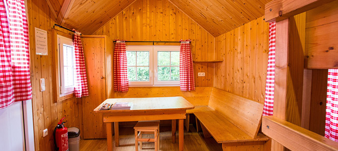 Kleine Holzhütte mit Tisch, Hockern, Sitzbank, rot karierten Vorhängen und einem Feuerlöscher in der Ecke.