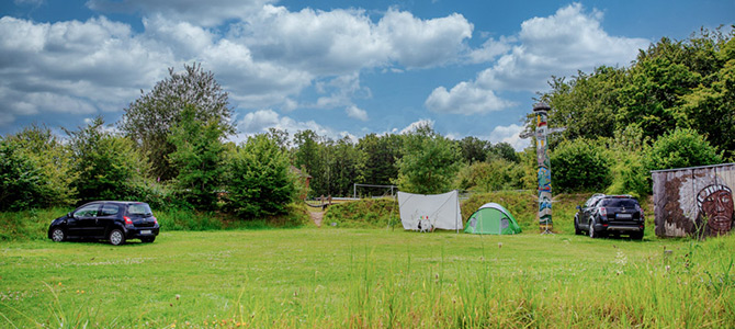 Twee auto's staan geparkeerd op een grasveld naast een witte luifel, een groene tent, een totempaal en een schuurtje met een muurschildering. Bomen en wolken zijn op de achtergrond te zien.
