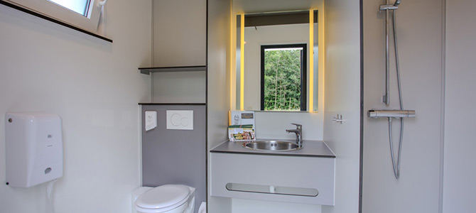 Modernes Badezimmer mit Wandtoilette, Waschbecken mit Spiegel und gelber Beleuchtung, Ablageflächen und einem Duschbereich mit Handbrause.
