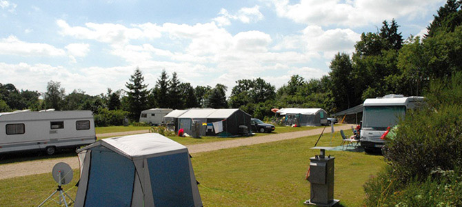 Ein Campingplatz mit Wohnwagen, Zelten und ein paar kleinen Gebäuden auf einer von Bäumen gesäumten Wiese unter einem teilweise bewölkten Himmel.