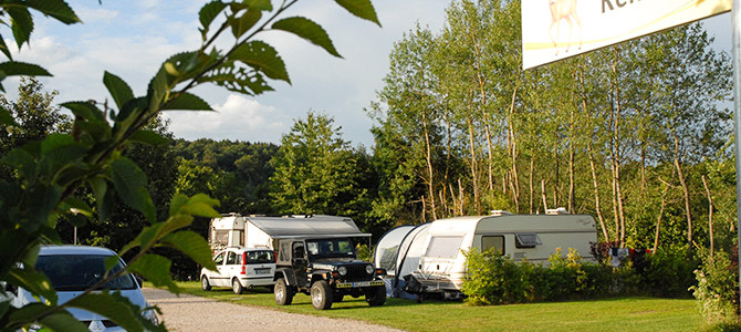 Mehrere geparkte Fahrzeuge und Wohnwagen stehen auf einer Schotterfläche neben Bäumen und Sträuchern, und im Vordergrund ist ein Teil des Campingplatzschildes zu sehen.