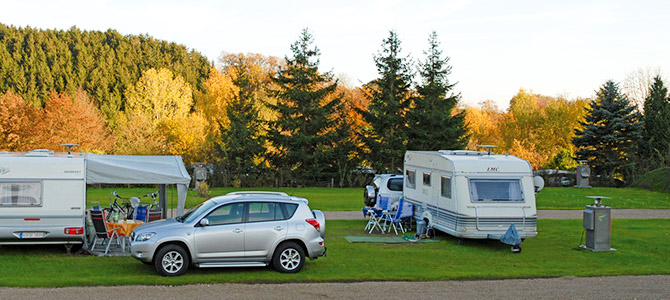 Zwei Wohnwagen und ein silberner Geländewagen stehen auf einem grasbewachsenen Campingplatz, auf dem Stühle und Tische im Freien aufgestellt sind. Im Hintergrund sind Bäume mit Herbstlaub zu sehen.