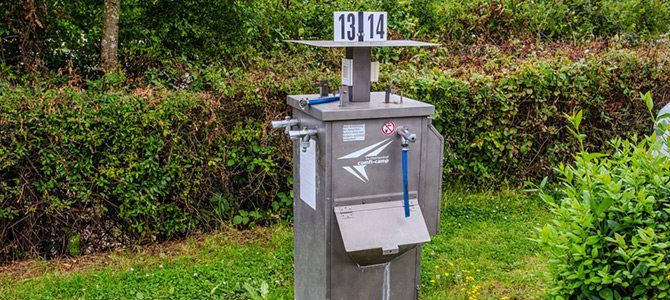 Eine Wasser- und Abfallentsorgungsstation aus rostfreiem Stahl mit nummerierten Schildern 13 und 14, die sich im Freien zwischen grünen Büschen befindet.