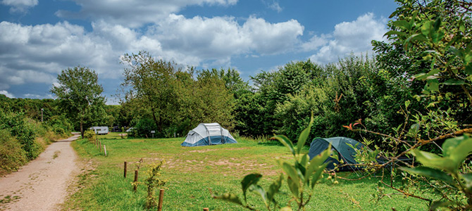 Een grascamping met twee opgezette tenten en een caravan in de verte, omringd door bomen onder een halfbewolkte hemel.