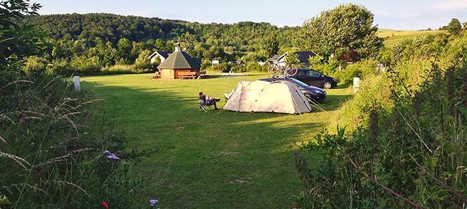 Een grasveld met een tent, auto's, een persoon in een stoel en een houten hut, omringd door bomen en heuvels op de achtergrond.
