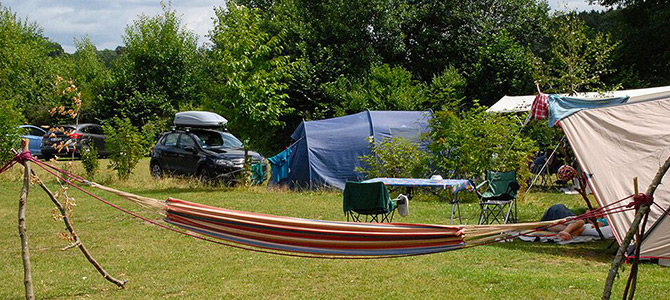Een gestreepte hangmat is vastgebonden tussen twee houten palen op een camping met tenten, klapstoelen, auto's en bomen op de achtergrond.